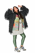 A woman modeling a black shaggy fringe jacket over a colorful graphic zip-up top, a light gray dress, metallic olive green leggings, and white sneakers, set against a plain white background.