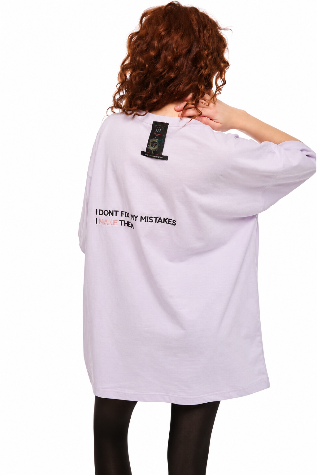 A model from behind wearing an oversized light lavender t-shirt with black text on the back reading 'I DON'T FOR MY MISTAKES I FORGET HER' and a black graphic patch near the neckline, paired with black tights against a white background.