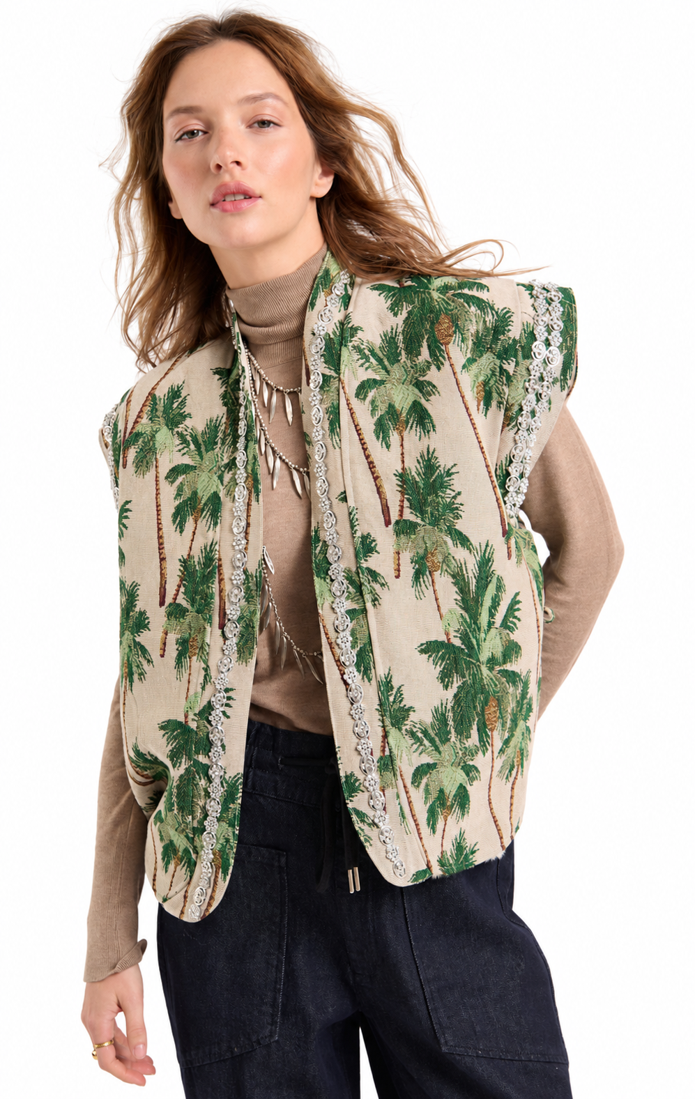 A woman modeling a sleeveless beige vest with a green palm tree print and silver beaded trim, worn over a brown turtleneck and dark pants against a white background.