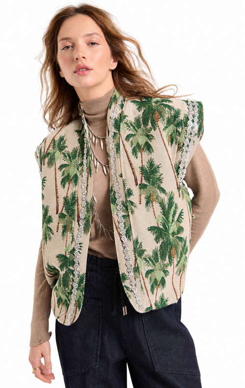 A woman modeling a sleeveless beige vest with a green palm tree print and silver beaded trim, worn over a brown turtleneck and dark pants against a white background.