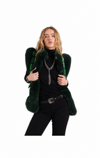 A model wearing a dark green faux fur vest with structured shoulders and green beaded fringe, styled with a black turtleneck, black pants, and silver jewelry against a plain white background.