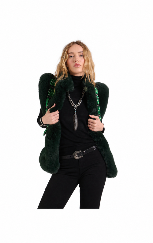 A model wearing a dark green faux fur vest with structured shoulders and green beaded fringe, styled with a black turtleneck, black pants, and silver jewelry against a plain white background.