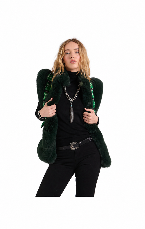 A model wearing a dark green faux fur vest with structured shoulders and green beaded fringe, styled with a black turtleneck, black pants, and silver jewelry against a plain white background.