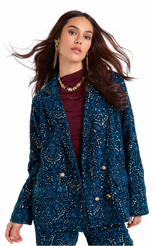 A woman modeling a dark teal blue double-breasted blazer with a gold geometric pattern, worn over a burgundy turtleneck. She is also wearing gold statement earrings and a gold chain necklace, set against a plain white background.