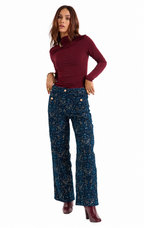 A woman modeling a dark burgundy asymmetrical long-sleeve top and dark blue patterned wide-leg pants with gold buttons, paired with burgundy heeled boots, against a plain white background.