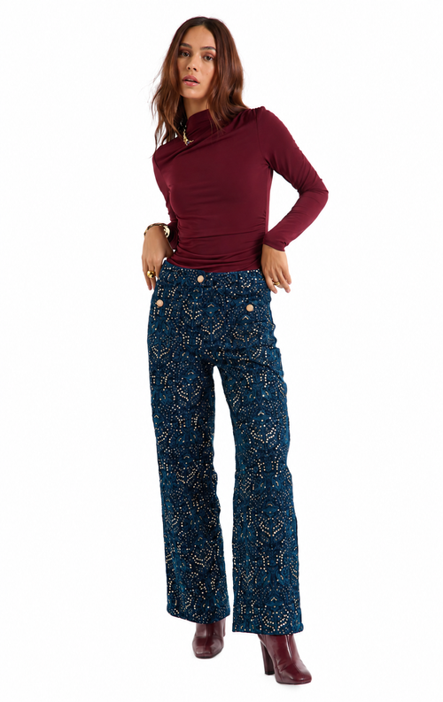 A woman modeling a dark burgundy asymmetrical long-sleeve top and dark blue patterned wide-leg pants with gold buttons, paired with burgundy heeled boots, against a plain white background.