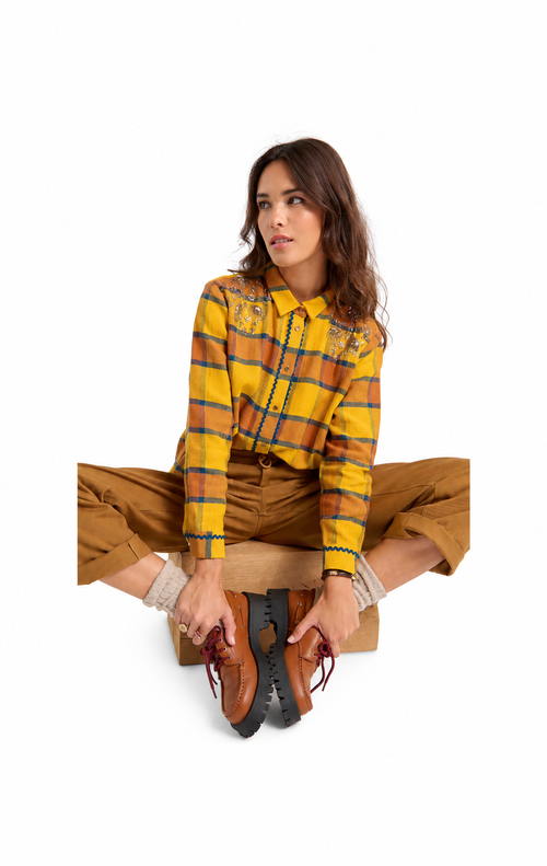 A woman sits cross-legged on a white studio background wearing a yellow, orange, and blue plaid long-sleeve western-style shirt, brown pants, and brown lace-up boots.