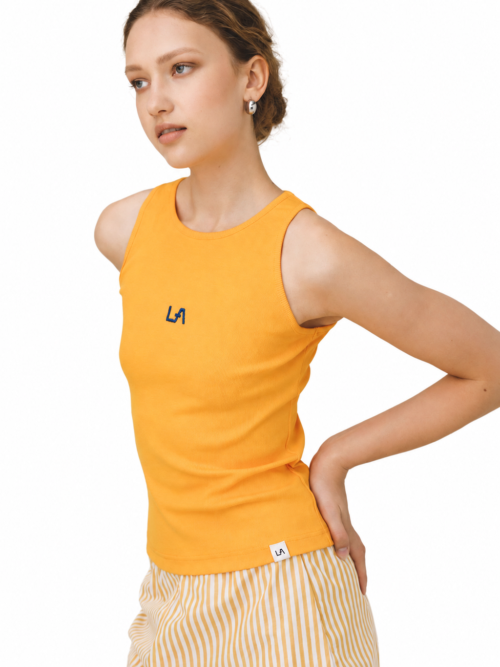 A woman modeling a bright yellow sleeveless tank top with a small navy 'LA' logo on the chest, paired with yellow and white striped pants, against a plain white background.