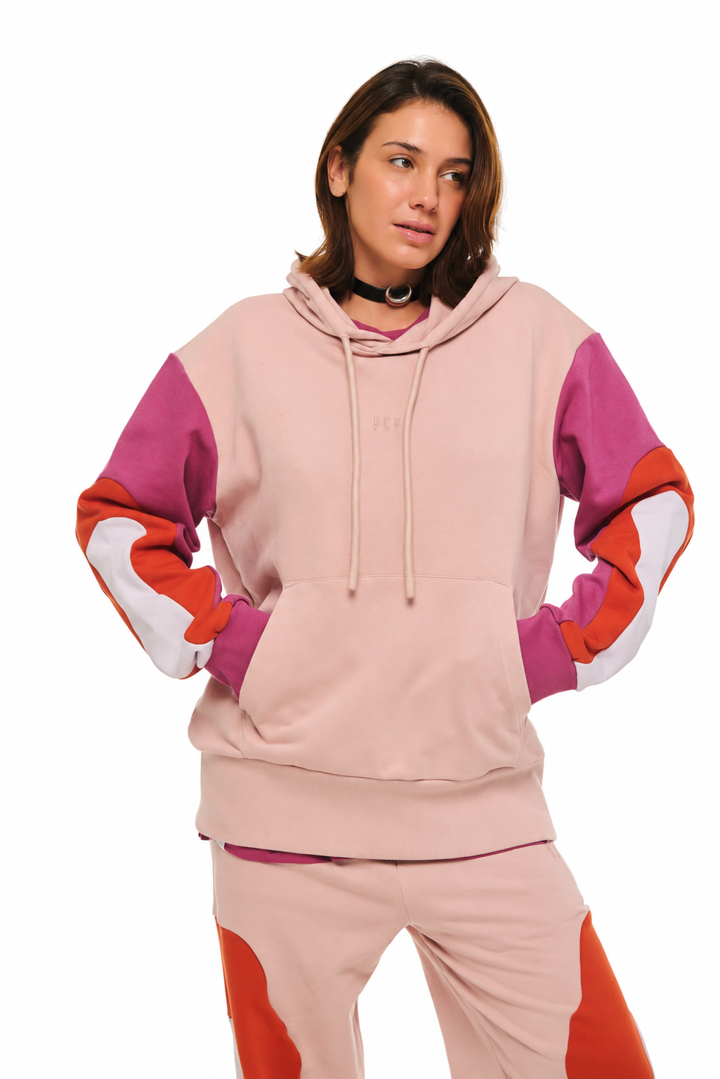 A model wearing a color-blocked hoodie and matching sweatpants. The hoodie features a light pink body with magenta, orange, and white color-blocked sleeves.