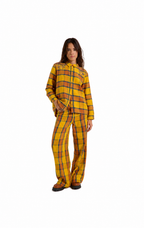 A woman modeling a matching two-piece plaid pajama set consisting of a long-sleeve button-front top and wide-leg pants, in a yellow, orange, and black tartan pattern, shot against a solid white background.