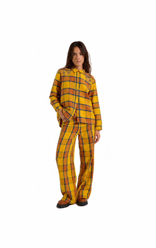 A woman modeling a matching two-piece plaid pajama set consisting of a long-sleeve button-front top and wide-leg pants, in a yellow, orange, and black tartan pattern, shot against a solid white background.
