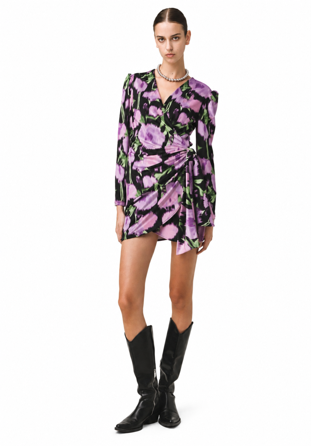 A model wearing a long-sleeve wrap mini dress with a purple and pink floral print on a black background, styled with black leather cowboy boots against a plain white studio background.