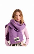 A woman wearing a fuzzy lavender-purple wrap scarf with fringe and a small white patch reading 'WILD' on the front, over a light pink top, against a plain white background.