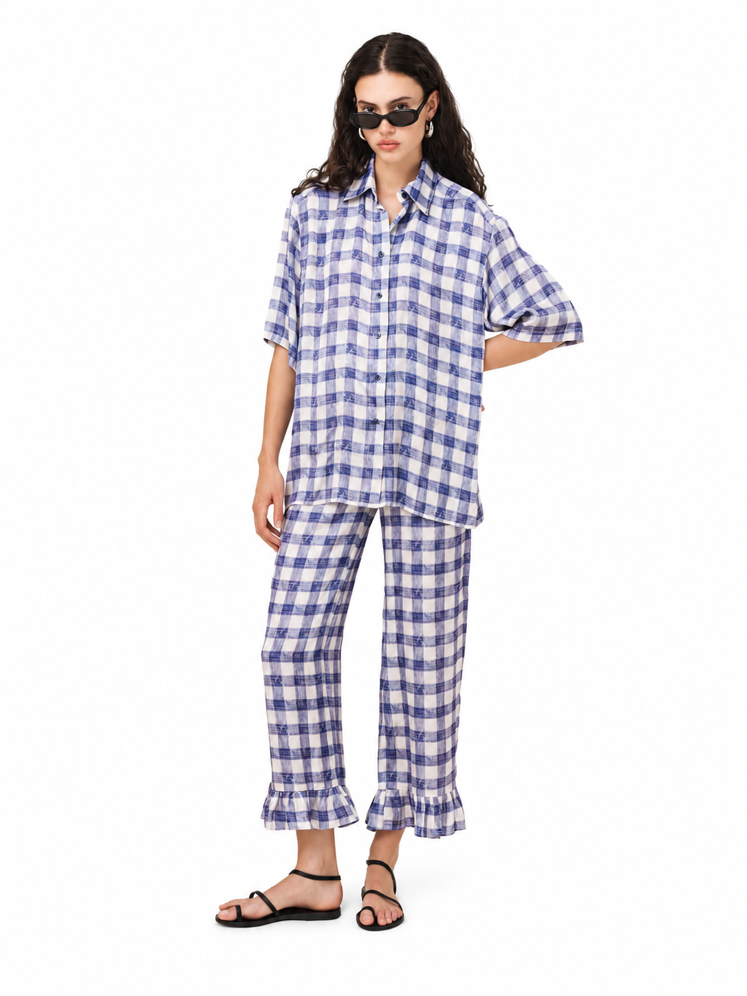 A model wearing a matching blue and white gingham check lounge set, consisting of a short-sleeve button-up shirt and ruffled ankle-length pants, styled with black strappy sandals against a plain white background.