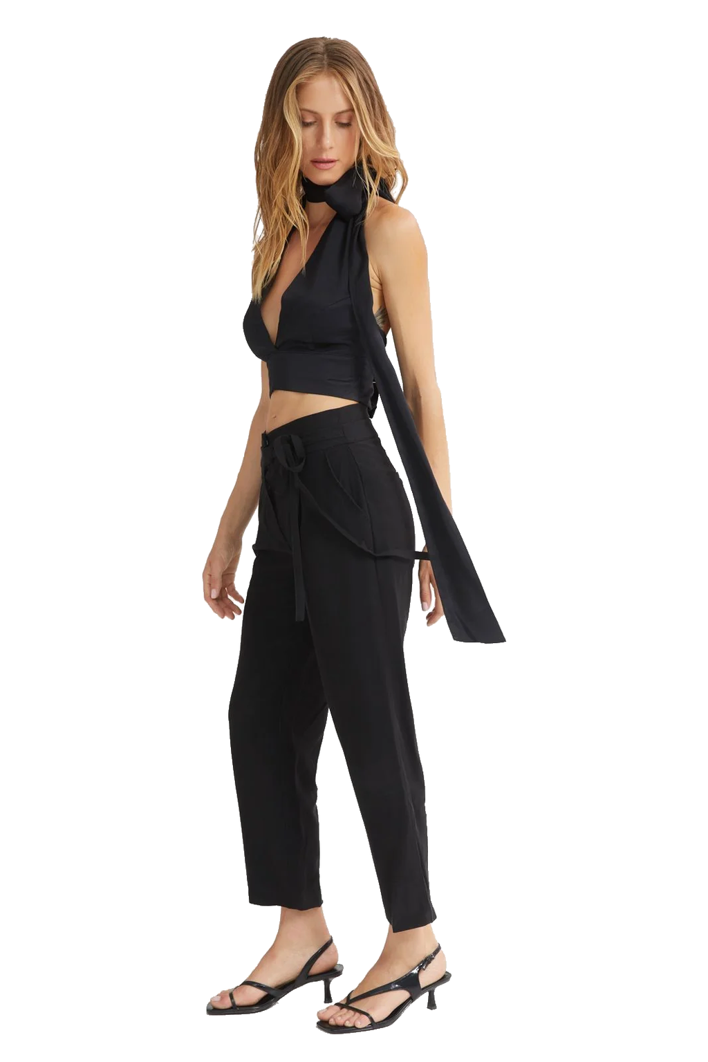 A woman modeling a black sleeveless halter crop top with a matching long draped tie detail, paired with black high-waisted cropped pants and black strappy heeled sandals, against a plain white background.