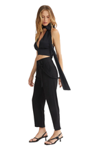 A woman modeling a black sleeveless halter crop top with a matching long draped tie detail, paired with black high-waisted cropped pants and black strappy heeled sandals, against a plain white background.