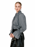 A model wearing a gray and black vertically striped long-sleeve blouse with a high collar and side tie, paired with black patent leather pants, against a plain white background.