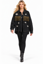 A woman modeling a black distressed denim jacket with gold fringe and embroidered details, paired with black pants and black boots, on a white studio background.