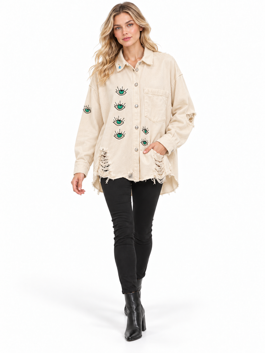 A woman modeling a distressed cream-colored denim jacket with green and black eye embroidery on the front, paired with black skinny jeans and black ankle boots, against a plain white background.
