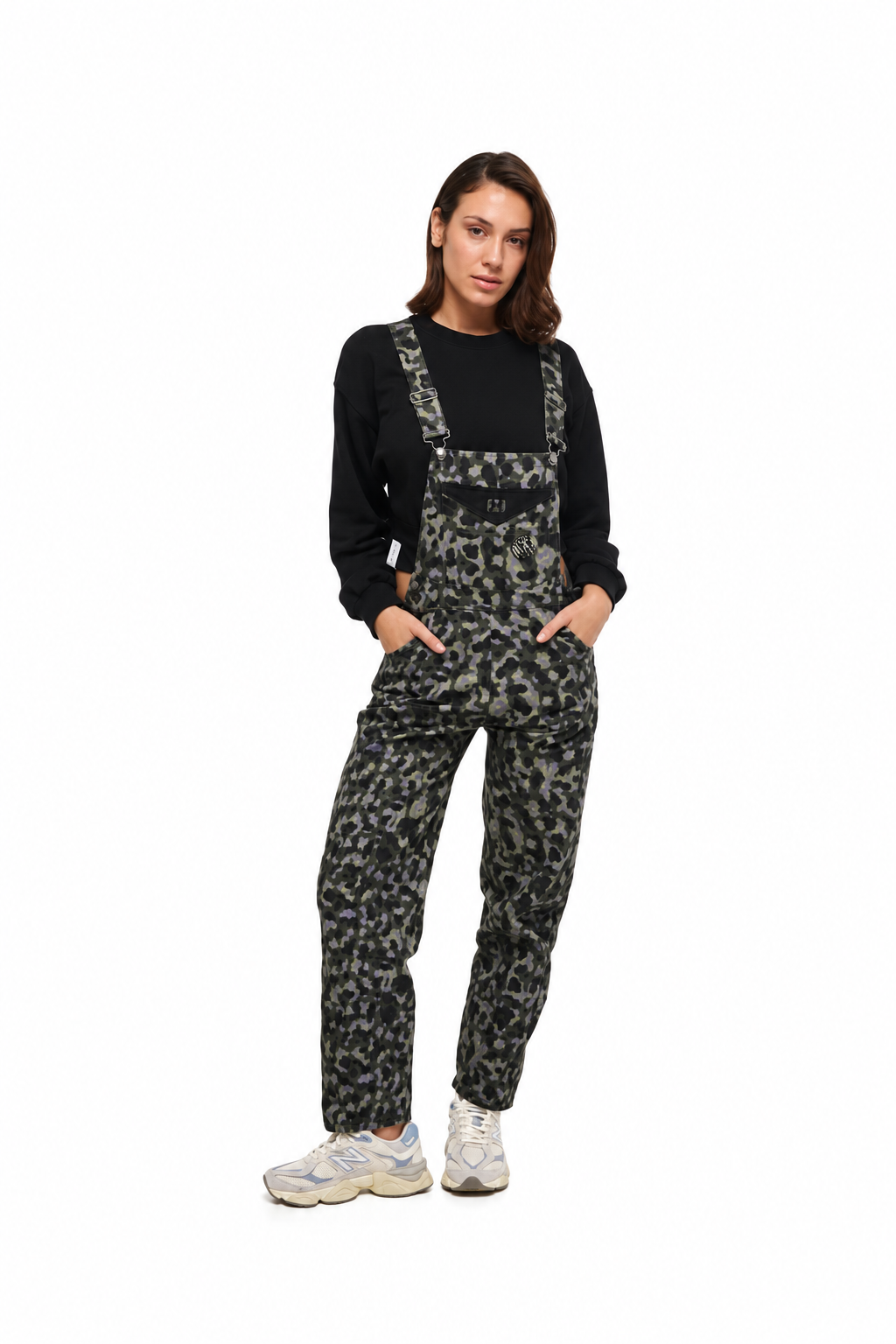 A woman modeling a pair of black and gray camouflage print overalls, worn over a black long-sleeve top and paired with white sneakers, shot against a solid white background.