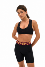 A woman modeling a black sports bra and matching black biker shorts with a burgundy waistband featuring gold 'PCP' lettering, set against a plain white background.