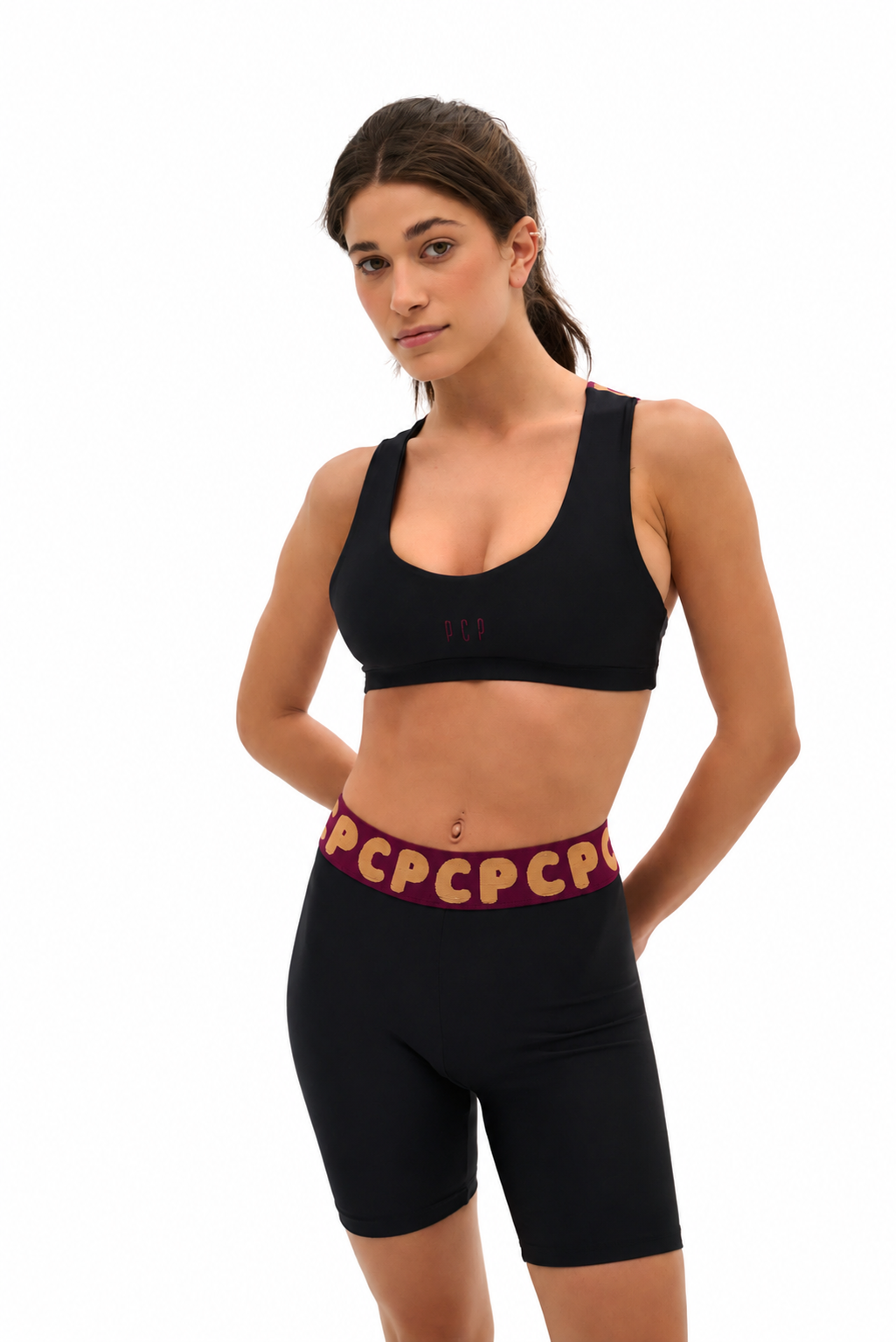 A woman modeling a black sports bra and matching black biker shorts with a burgundy waistband featuring gold 'PCP' lettering, set against a plain white background.