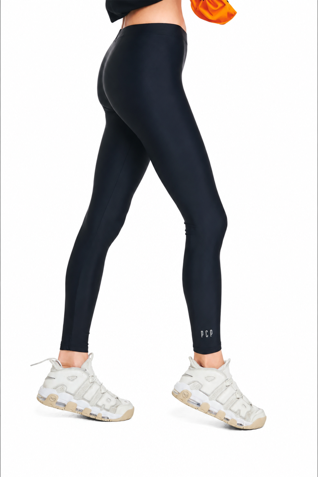 A model from the waist down wearing black high-waisted leggings with a small white logo on the lower left leg, paired with white sneakers, against a plain white background.