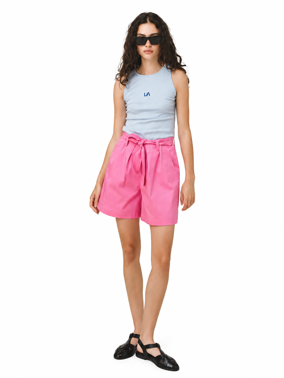A model wearing a light blue sleeveless top with a small blue LA logo, bright pink high-waisted shorts with a tie waist, and black flat shoes, photographed against a solid white background.