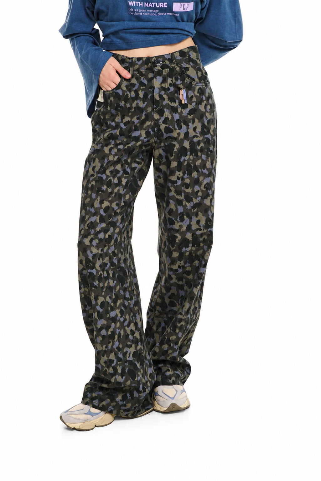 A person wearing loose-fit camouflage pants in olive green, black, and gray, paired with a blue cropped sweatshirt and beige sneakers, shown against a plain white background.