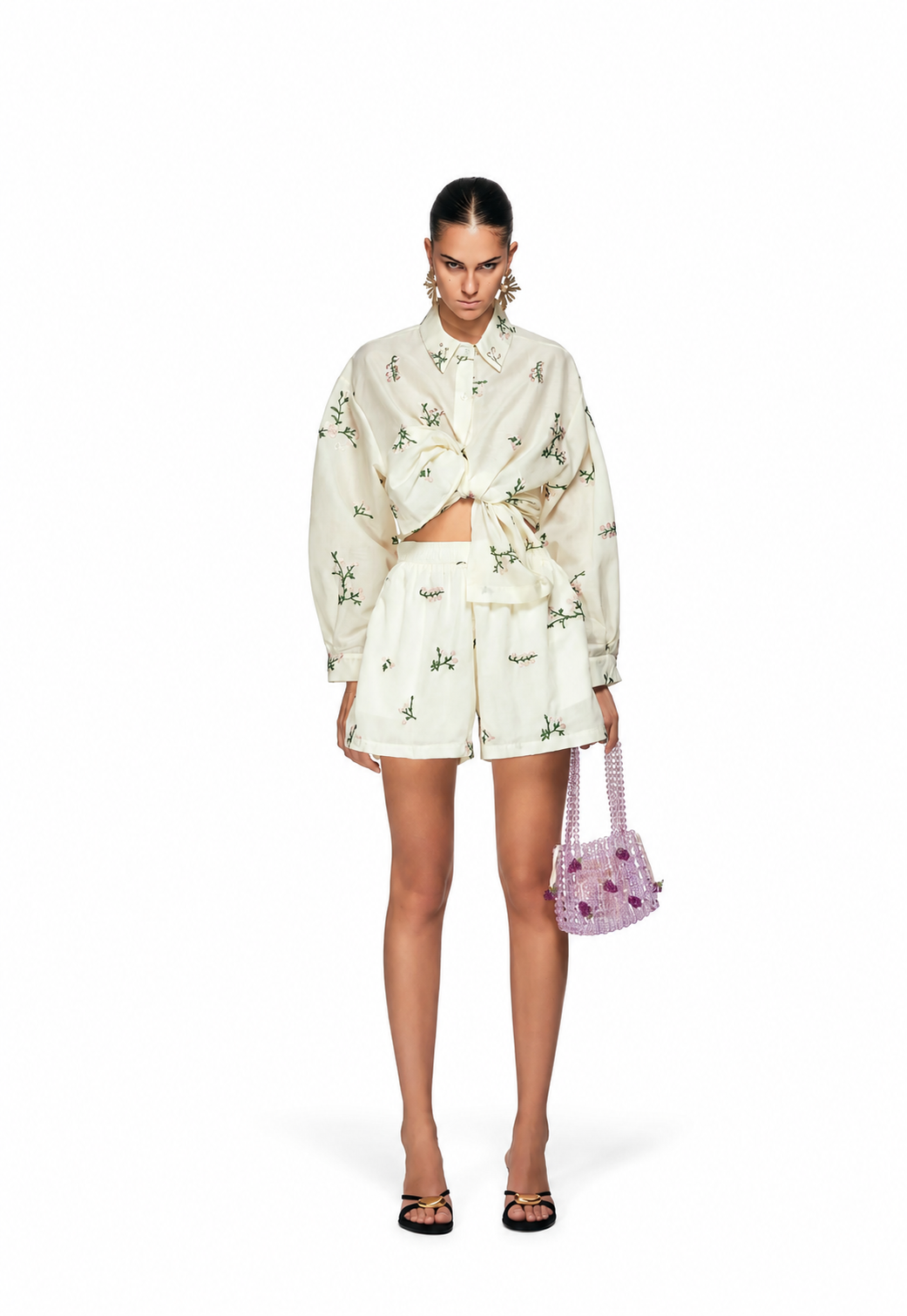 A model stands against a white studio background wearing a cream-colored two-piece set with a green floral print, consisting of a long-sleeve cropped tie-front shirt and matching shorts. She is holding a small pink floral handbag and wearing black flat sandals.