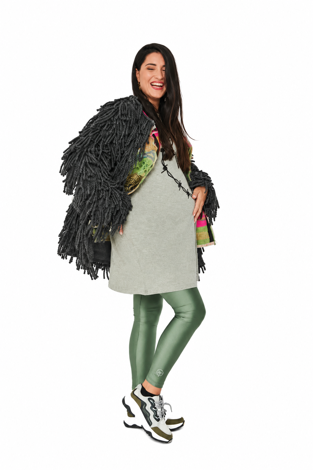 A pregnant woman posing in a studio setting, wearing a light gray maternity dress, metallic olive green leggings, a black shaggy fringe cardigan, and chunky white and olive sneakers. She is holding a colorful patterned clutch bag against a solid white background.