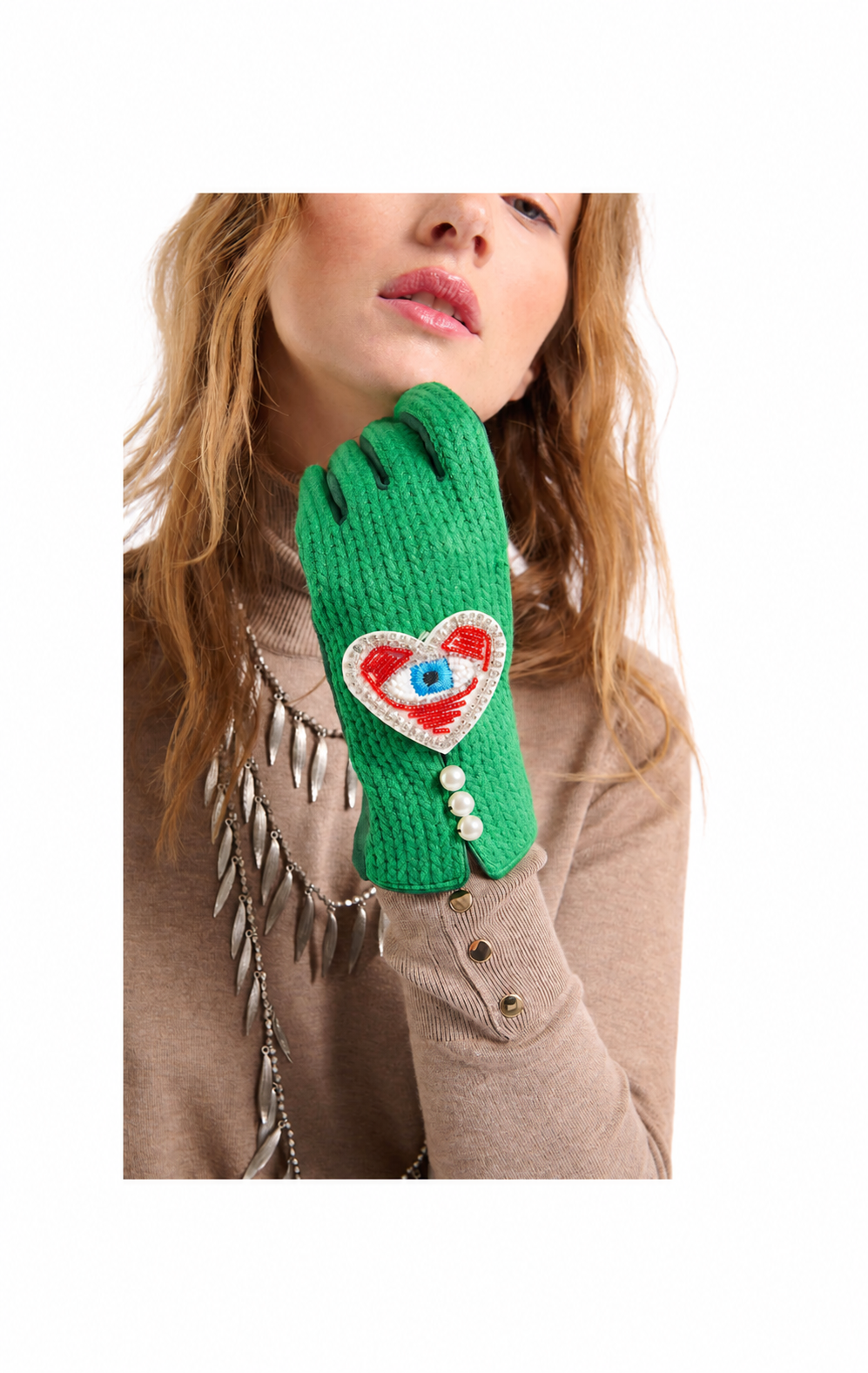 A close-up of a model wearing a bright green knit glove with a red and blue heart patch featuring an eye design, paired with a beige sweater.