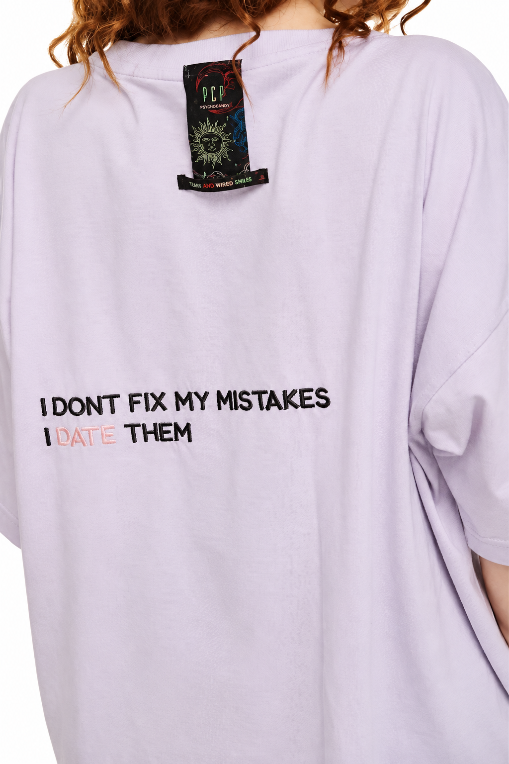 A close-up of a person wearing an oversized light lavender t-shirt. The back of the shirt features the printed text 'I DONT FIX MY MISTAKES I DATE THEM' in black and orange-red.