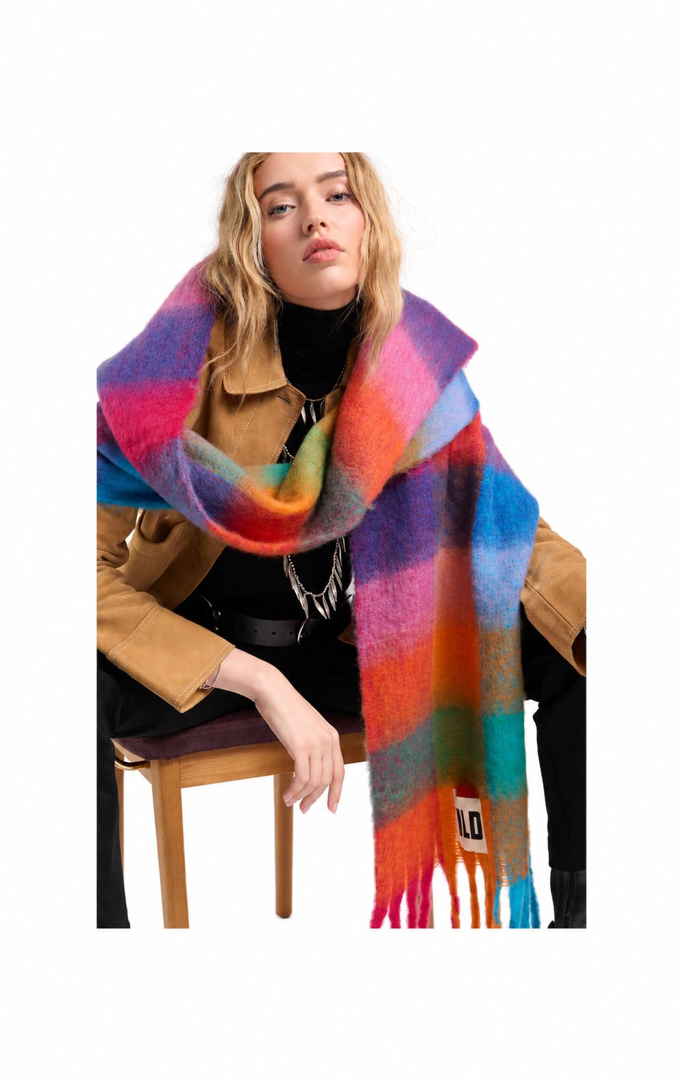 A model wearing a vibrant, multicolored striped fuzzy scarf with fringe, styled over a tan jacket and black top, seated on a wooden chair against a white background.