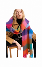 A model wearing a vibrant, multicolored striped fuzzy scarf with fringe, styled over a tan jacket and black top, seated on a wooden chair against a white background.