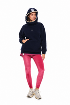 A model wearing a navy blue fleece hoodie with a white embroidered logo, paired with bright pink leggings and white sneakers, standing against a solid white background.