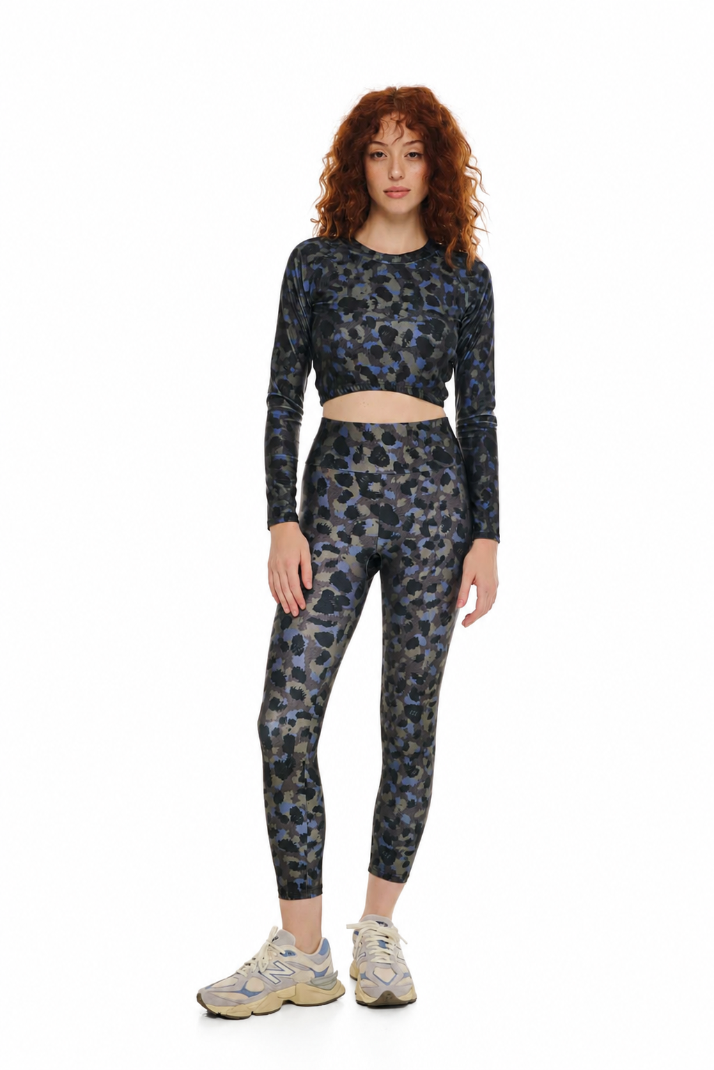 A model with curly red hair wearing a matching two-piece activewear set featuring a blue, gray, and black leopard print. The set includes a long-sleeve crop top and high-waisted leggings, styled with beige and gray sneakers against a plain white background.