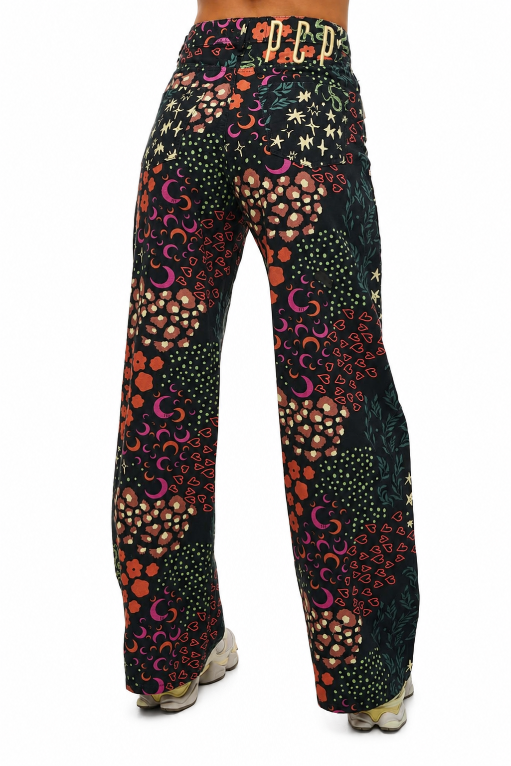 A pair of wide-leg pants with a complex psychedelic pattern featuring crescent moons, stars, dots, and floral motifs in orange, pink, yellow, and white on a black background, shown from the back on a model.