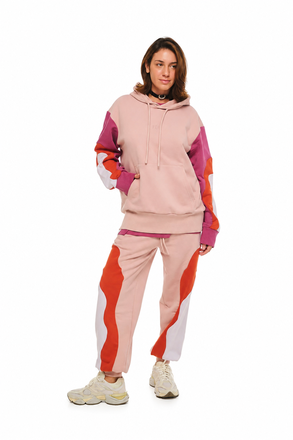 A woman modeling a two-piece color-blocked tracksuit featuring a light pink hoodie and matching joggers with purple, orange, and white color-blocked panels, set against a plain white background.