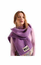 A woman wearing a fuzzy lavender-purple scarf with fringe and a small beige patch that reads 'WILD', paired with a light pink top against a plain white background.