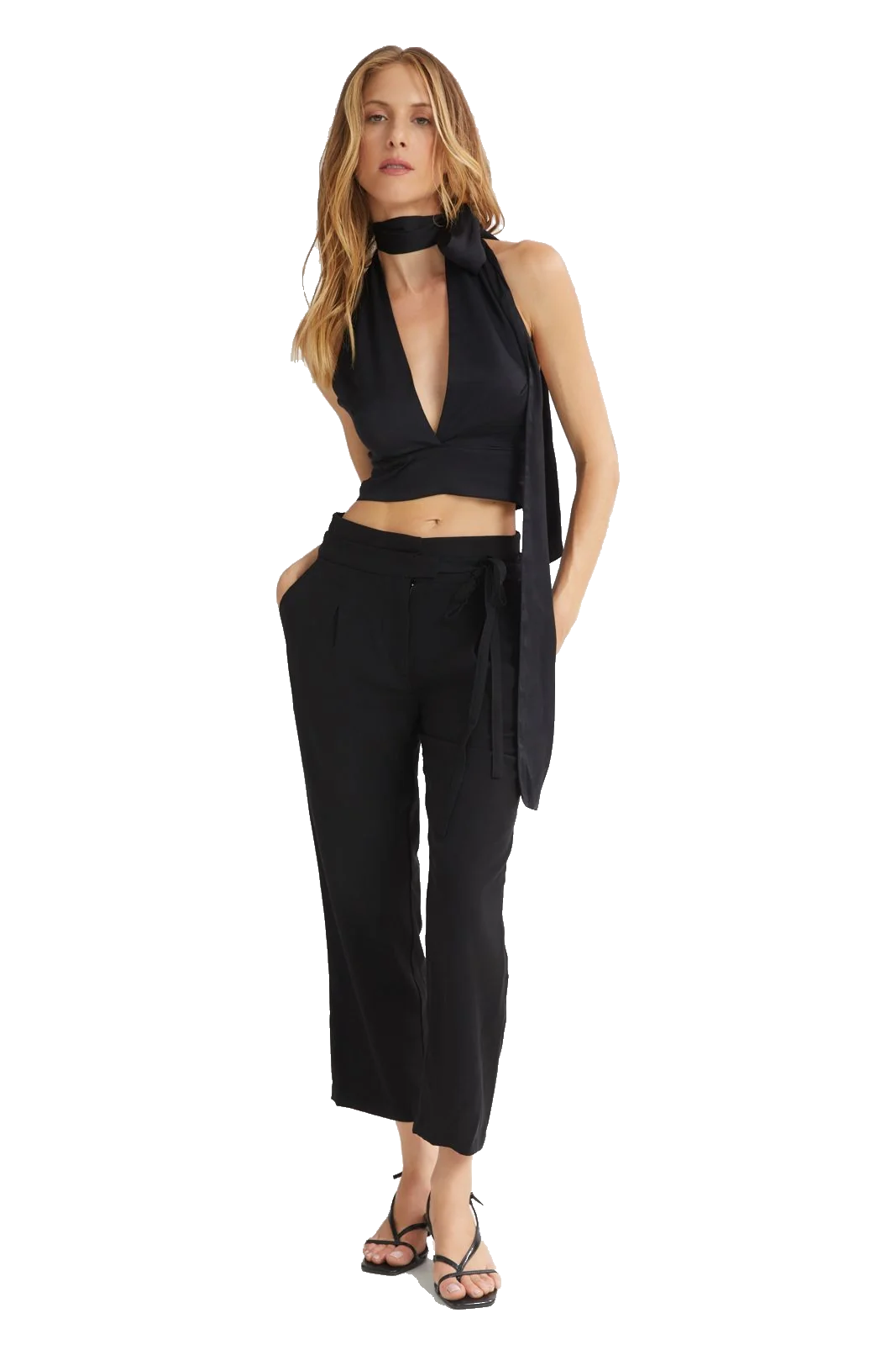 A woman modeling a black two-piece outfit consisting of a halter crop top with a long side drape and matching high-waisted cropped pants, paired with black strappy sandals against a plain white background.