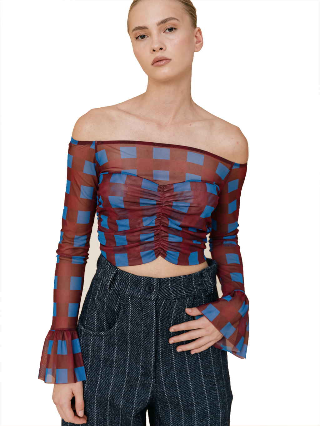 A model wearing a sheer off-the-shoulder long-sleeve crop top with a blue and burgundy checkered pattern, paired with dark pinstriped trousers, against a plain white background.