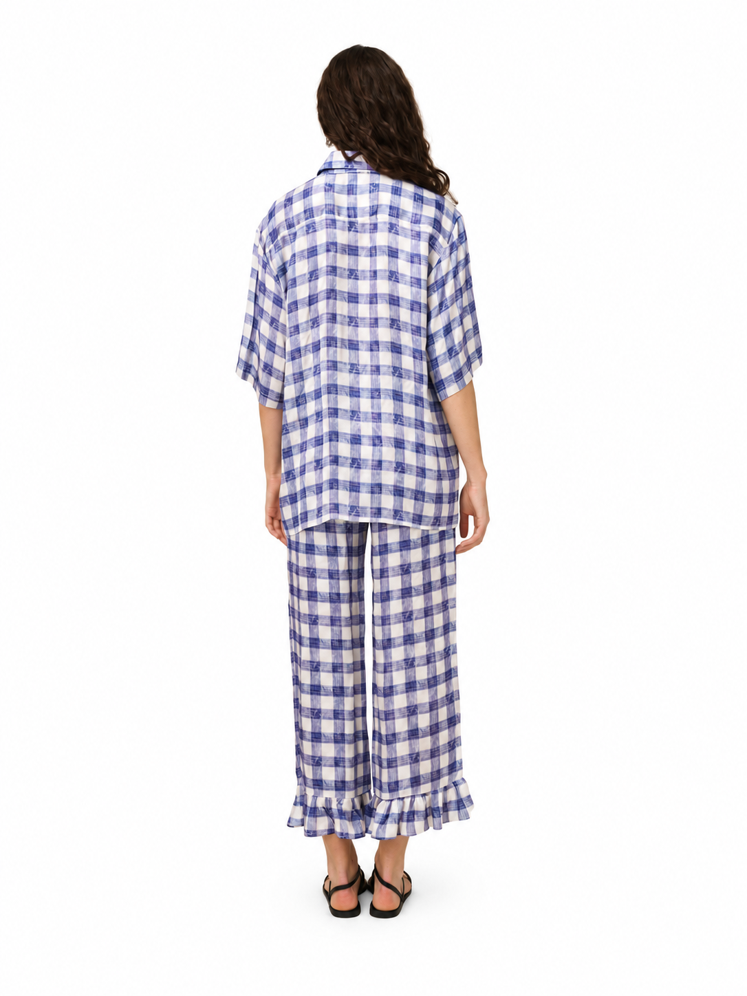 A woman viewed from behind wearing a matching blue and white gingham pajama set with a short-sleeve button-up shirt and wide-leg pants with ruffled hems, paired with black sandals, on a solid white background.
