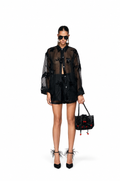 A model stands against a white studio background wearing a sheer black long-sleeve button-up shirt with floral embroidery, black shorts, black high heels with feather details, and holding a black embellished handbag.