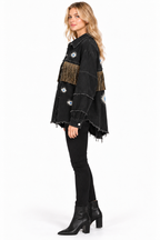 A woman modeling a black fringed denim jacket with embroidered details, paired with black skinny jeans and black ankle boots, against a plain white background.