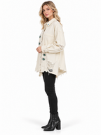 A woman modeling a distressed off-white denim jacket with green and blue embroidered details, paired with black skinny jeans and black ankle boots, against a plain white background.