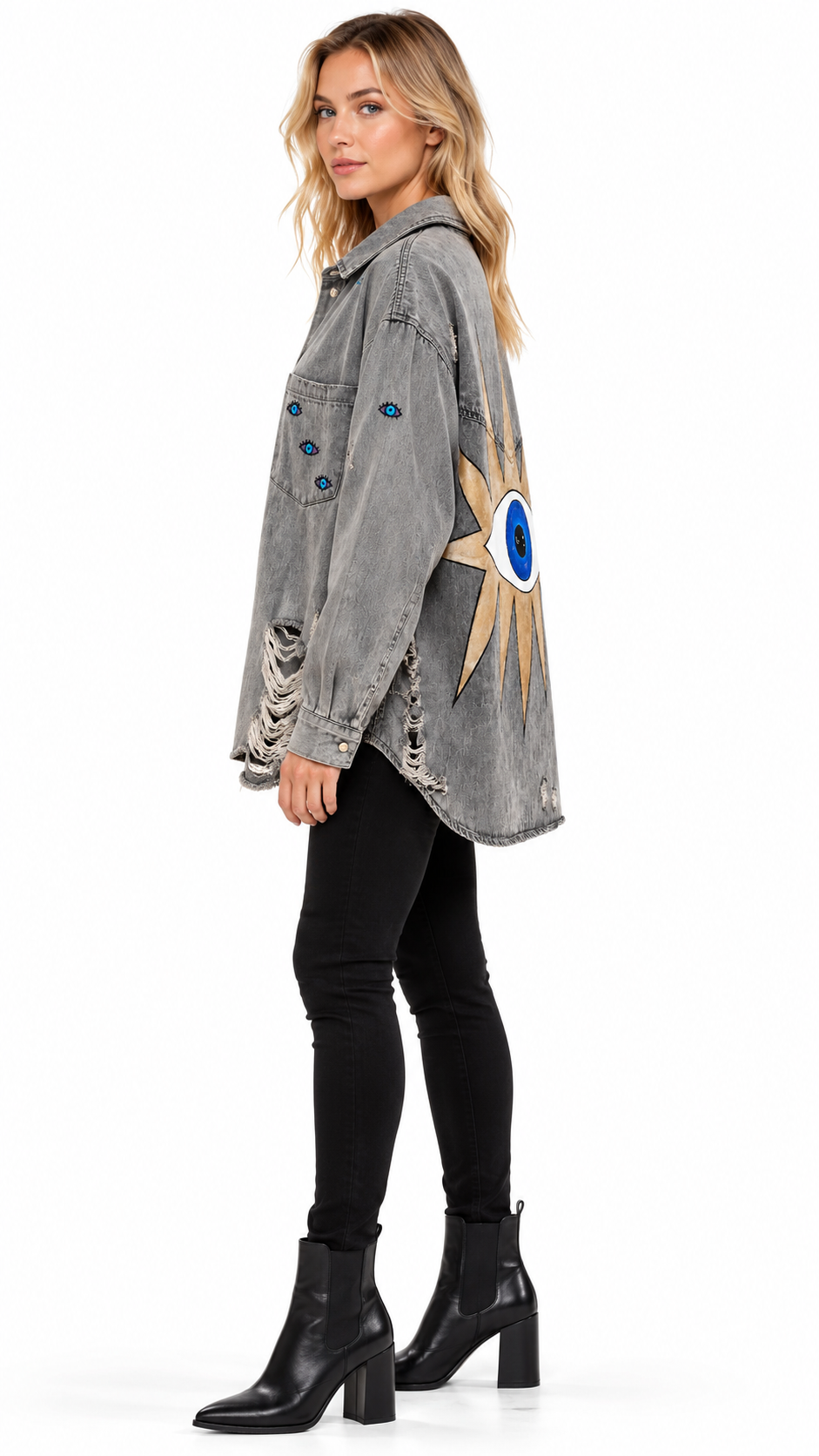 A woman modeling a washed gray denim jacket with a large sun and evil eye graphic on the sleeve, paired with black pants and black ankle boots against a white background.