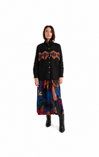 A model stands against a white studio background wearing a black button-front jacket with a colorful geometric chest pattern, a long multicolor floral skirt, and black boots.