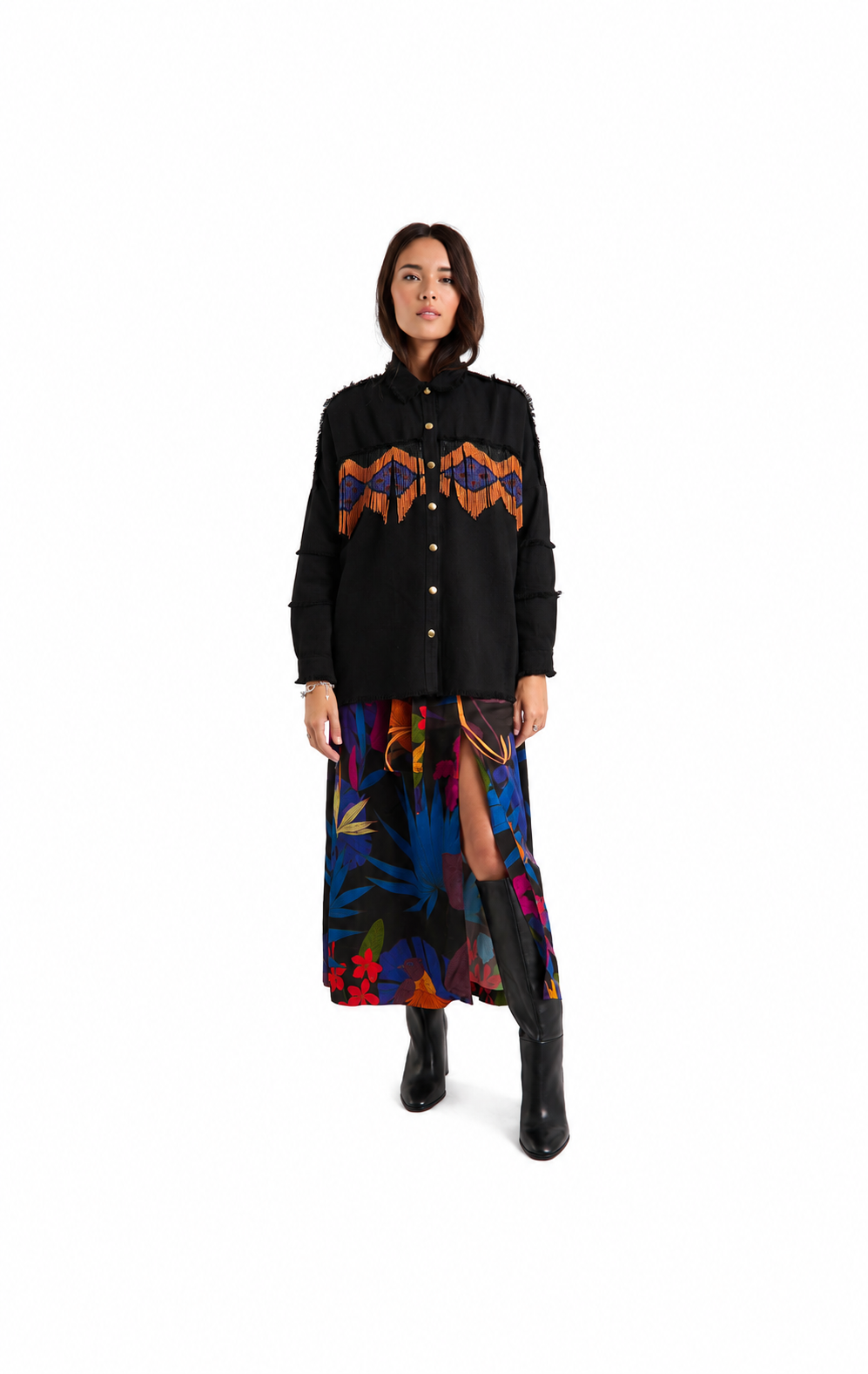 A model stands against a white studio background wearing a black button-front jacket with a colorful geometric chest pattern, a long multicolor floral skirt, and black boots.