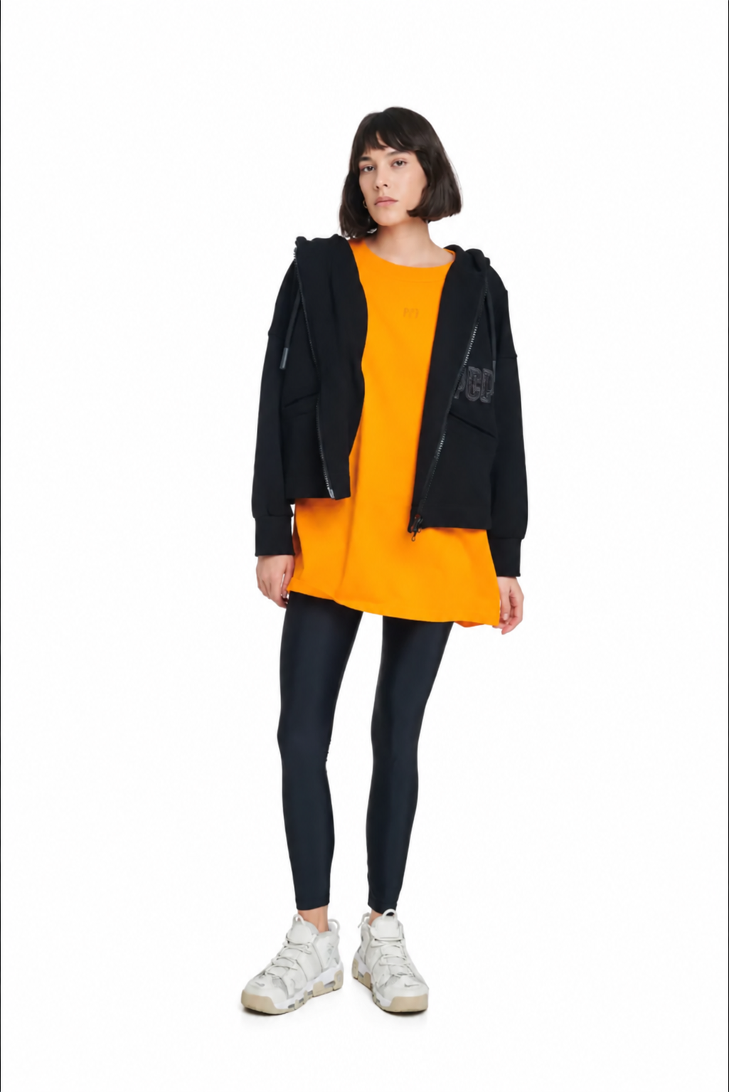 A model wearing a black zip-up hoodie over a bright orange tunic, paired with black leggings and white sneakers, standing against a plain white background.